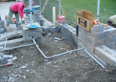 Workers constructing a brick structure with visible PVC pipes and scattered materials on the ground. A box and construction tools are nearby.